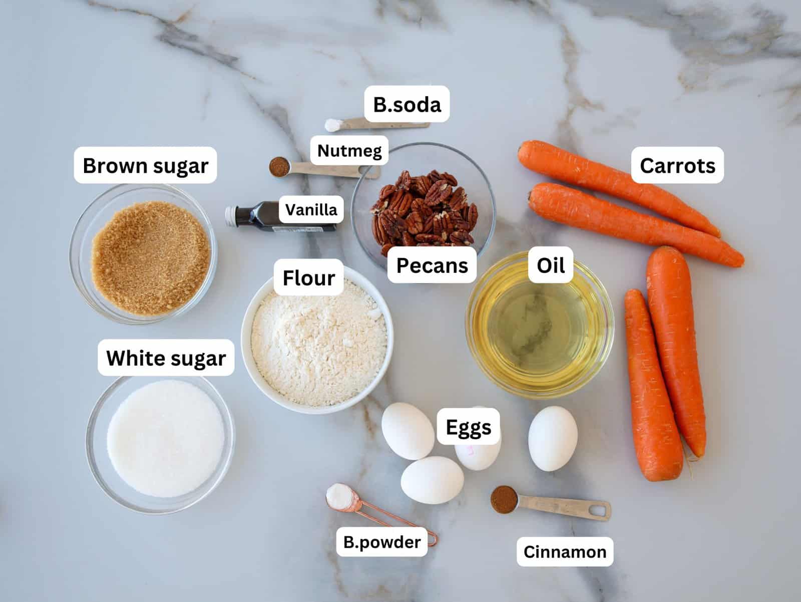 Baking ingredients on a marble surface labeled: brown sugar, white sugar, flour, baking powder, baking soda, nutmeg, vanilla, pecans, oil, eggs, cinnamon, and whole carrots.