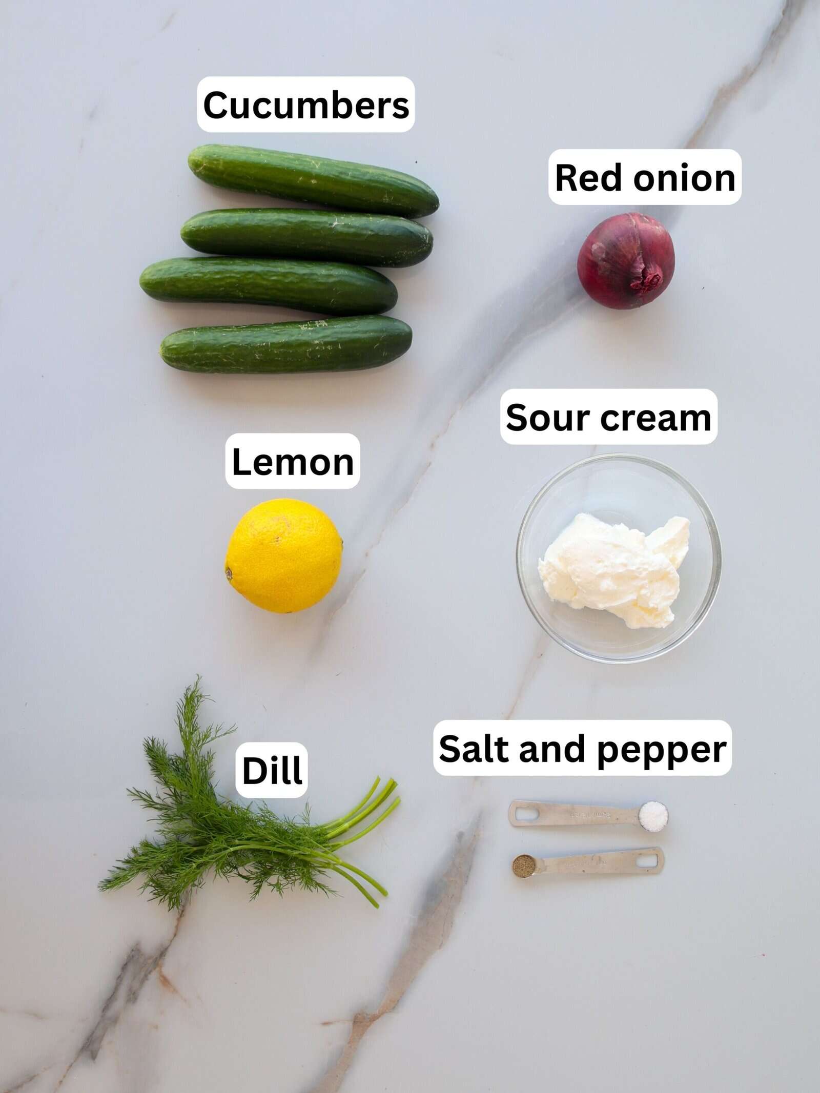 Five cucumbers, a red onion, a lemon, a bunch of dill, a bowl of sour cream, and measuring spoons of salt and pepper are arranged on a white marble surface, each labeled with text.