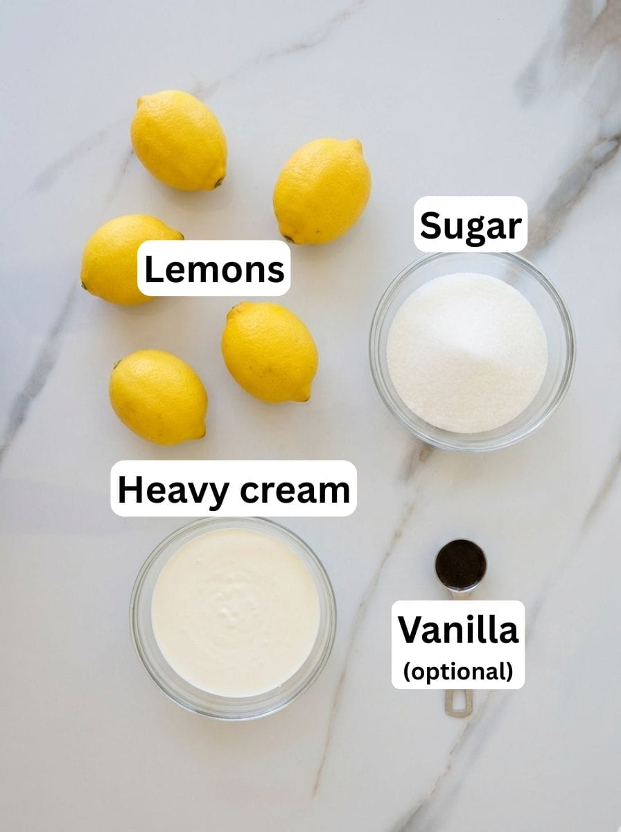 Five lemons, a bowl of sugar, a bowl of heavy cream, and a small dish of vanilla bean paste (optional) are arranged on a white marble surface, each labeled accordingly.