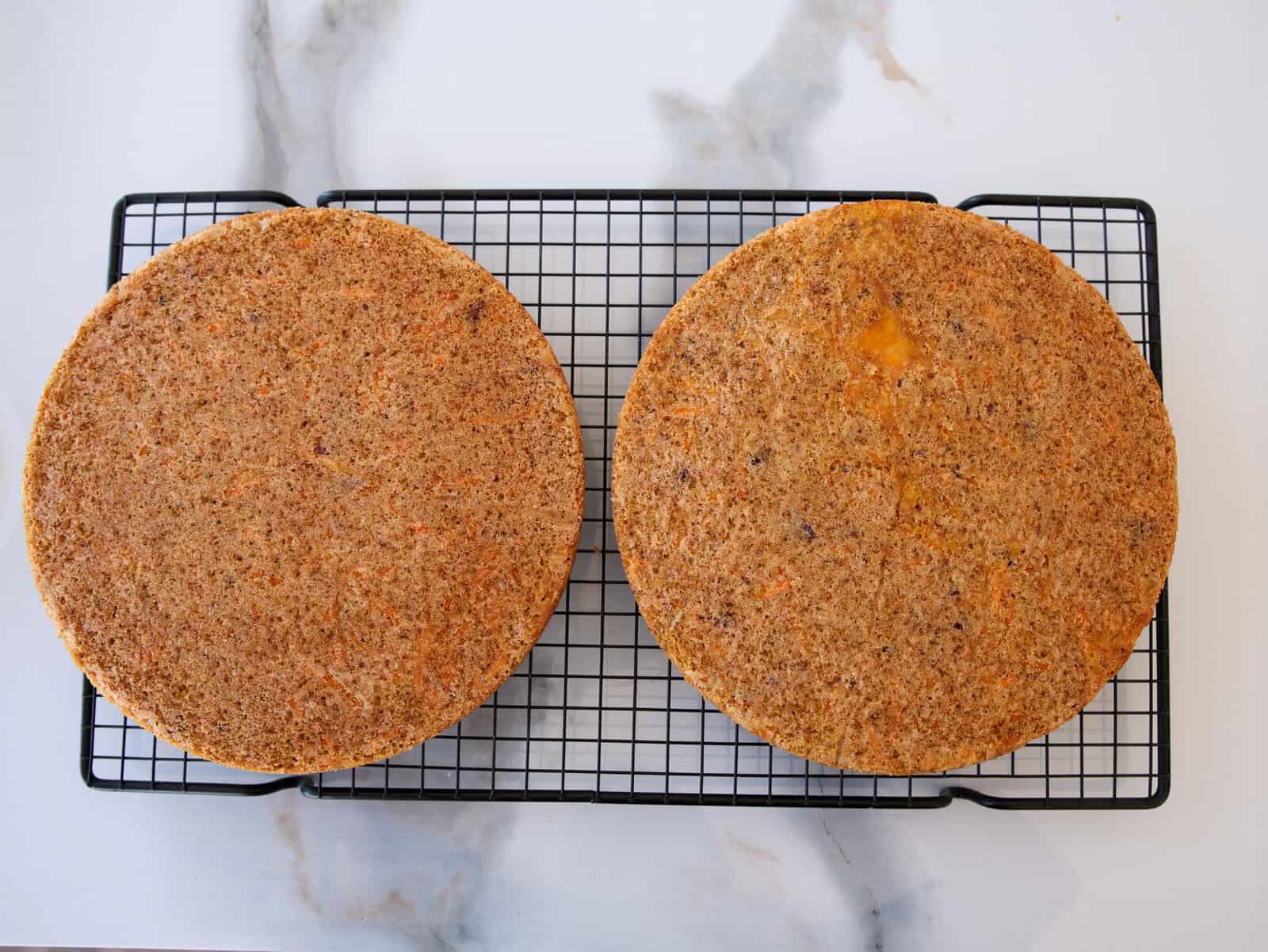 Two round, golden-brown cake layers are cooling on a black wire rack placed on a white marble surface. The cakes have a slightly textured surface.