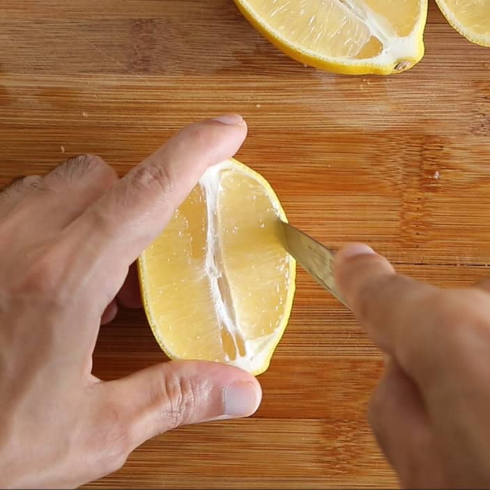 A person cuts a lemon half with a knife on a wooden cutting board. Another lemon half is visible at the top of the image.