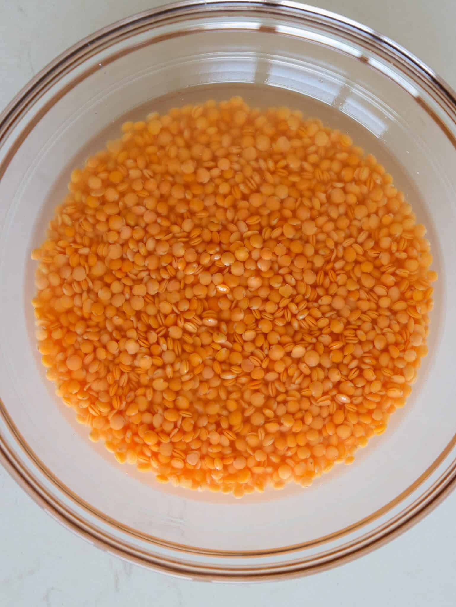 A clear glass bowl filled with water and soaked orange lentils, placed on a light-colored surface.