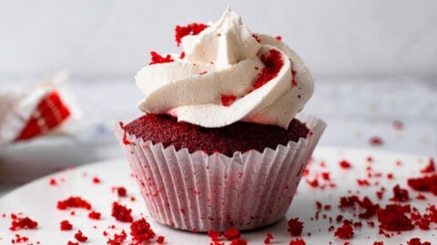A red velvet cupcake with white frosting sits on a plate, topped with red crumbs. Red cake crumbs are scattered around the cupcake on the white surface.