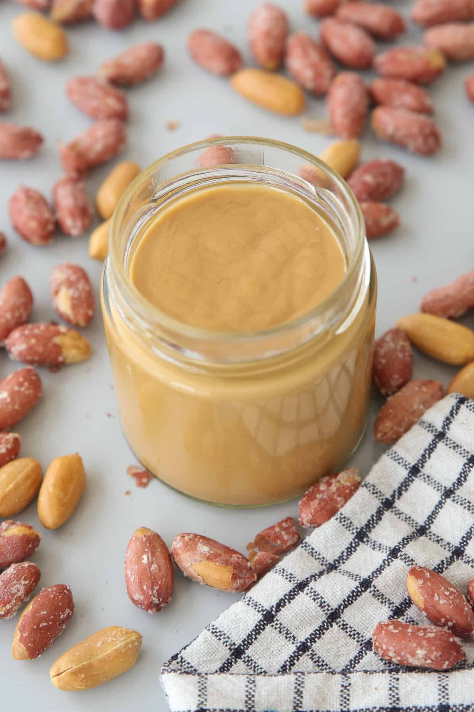 A glass jar filled with smooth peanut butter sits on a gray surface surrounded by shelled and unshelled peanuts. A black and white checkered cloth is placed nearby.