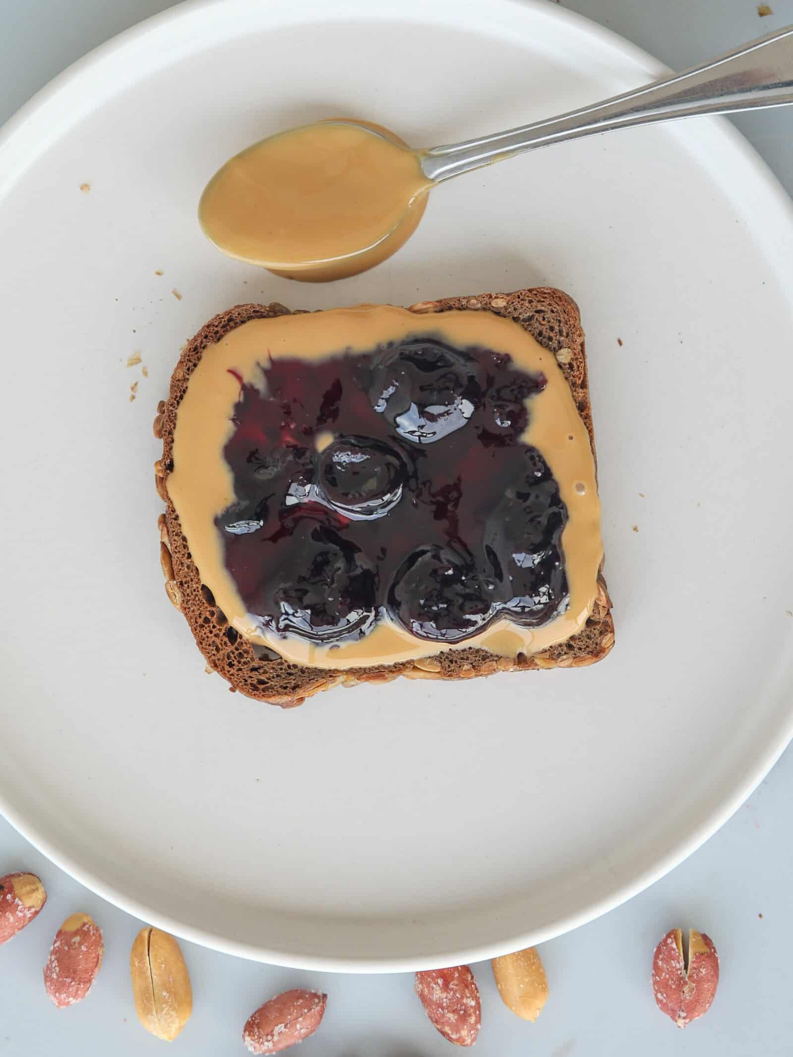 A slice of toast with peanut butter and blueberry jam on a white plate, a spoon with peanut butter above it, and peanuts arranged along the plate’s edge.