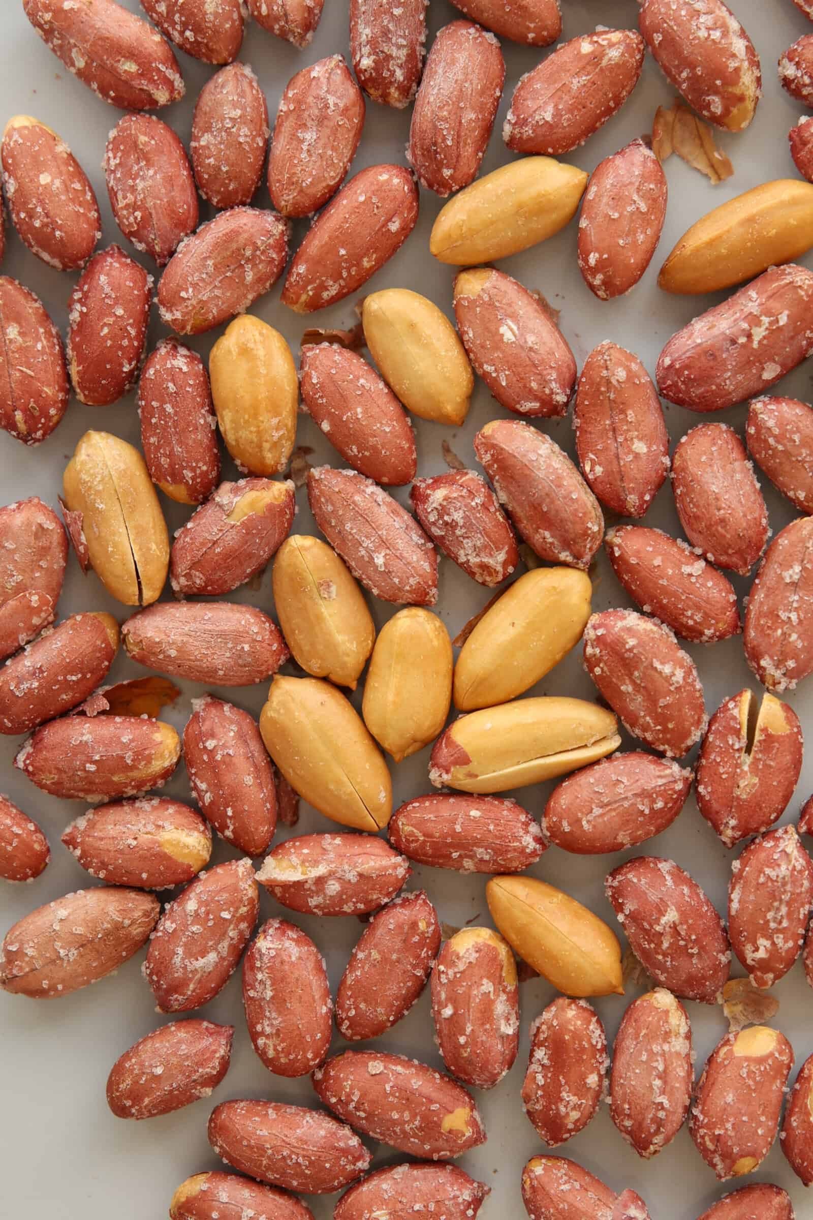 A close-up view of roasted peanuts, some with reddish-brown skins and others without skins, scattered on a light gray surface. Salt crystals are visible on the nuts.