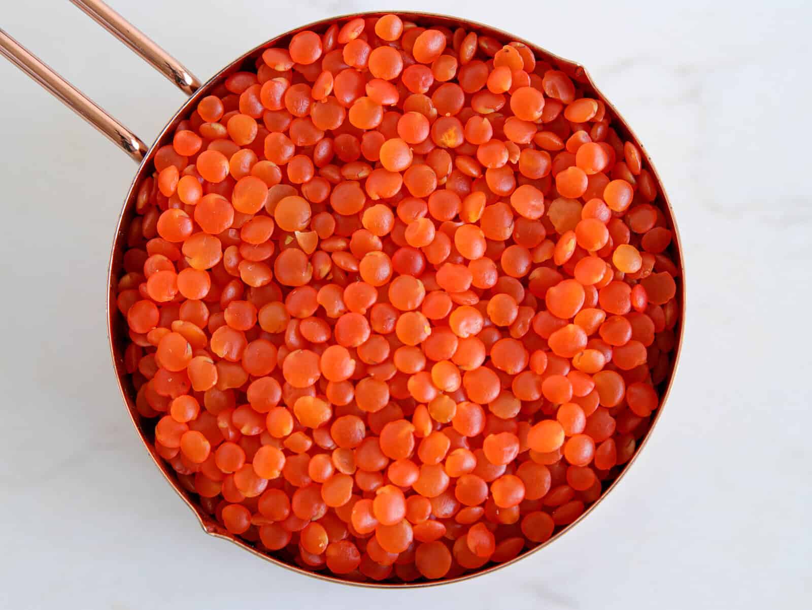 A copper measuring cup filled to the top with dry red lentils, viewed from above on a white surface.