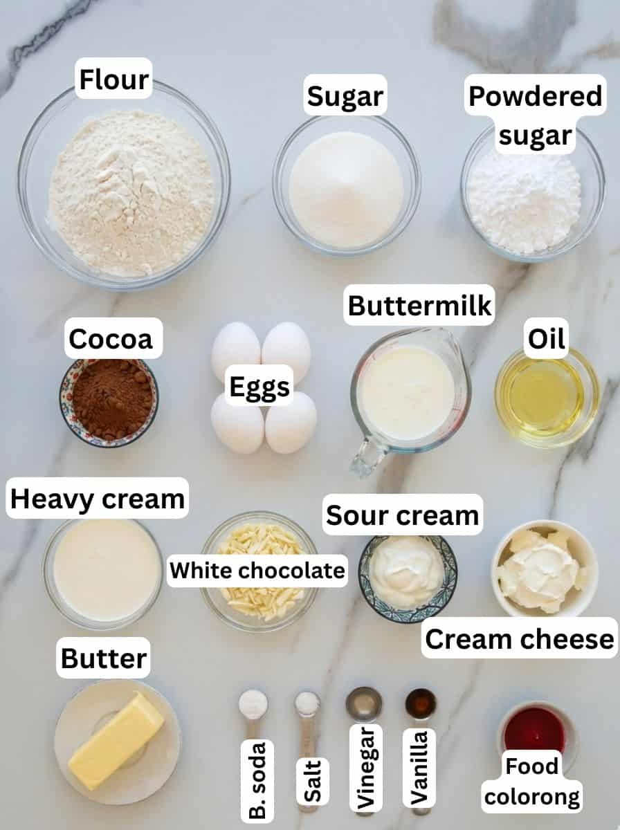 A variety of baking ingredients arranged on a white surface, each labeled: flour, sugar, powdered sugar, cocoa, eggs, buttermilk, oil, heavy cream, sour cream, white chocolate, cream cheese, butter, baking soda, salt, vinegar, vanilla, and food coloring.