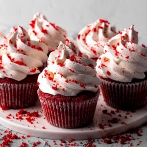 Five red velvet cupcakes with generous swirls of white frosting, topped and surrounded by red cake crumbs, are displayed on a white plate against a light background.