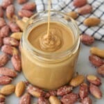 A stream of creamy peanut butter is being poured into an open glass jar, surrounded by shelled and unshelled peanuts on a gray surface with a checkered cloth in the background.
