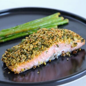 A piece of herb-crusted salmon with a crunchy topping is served on a black plate alongside a small bundle of cooked asparagus spears.