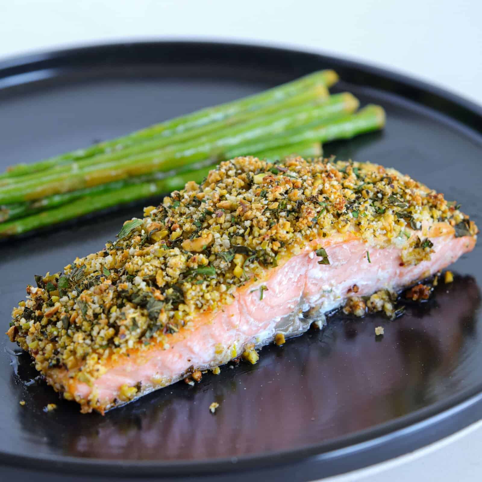 A piece of herb-crusted salmon with a crunchy topping is served on a black plate alongside a small bundle of cooked asparagus spears.
