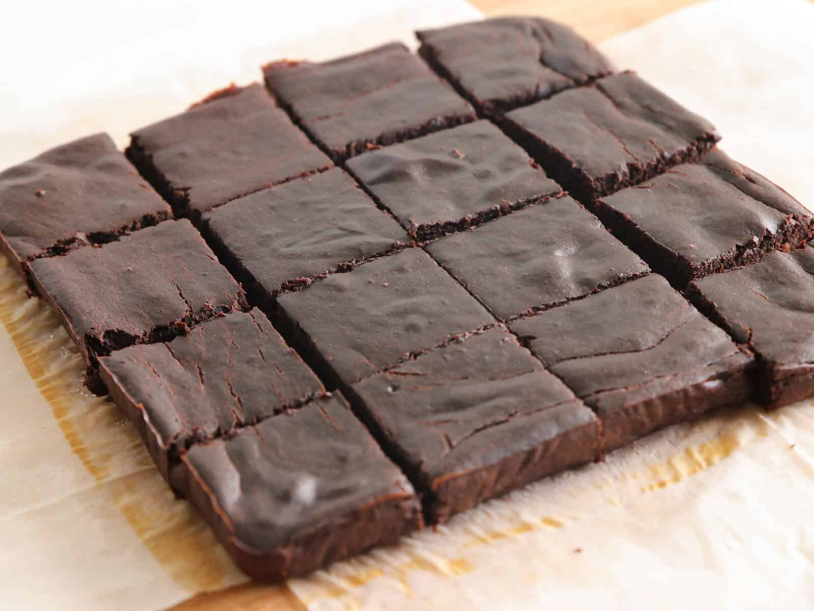 A batch of healthy chocolate brownies cut into sixteen squares, resting on parchment paper on a wooden surface.