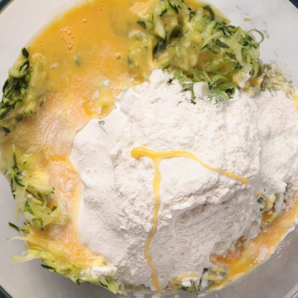 A glass bowl containing shredded zucchini, flour, and beaten eggs, ready to be mixed for a recipe. The ingredients are unmixed, with the flour piled in the center.