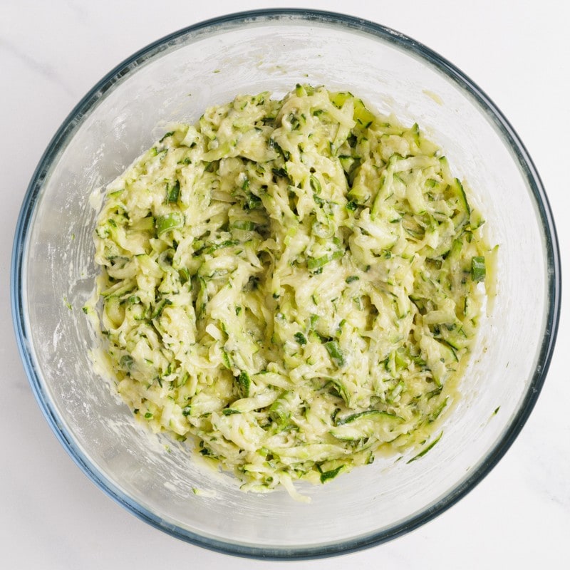 A clear glass bowl filled with a mixture of shredded zucchini, flour, and spices, forming a thick, light green batter. The bowl is set on a white surface.