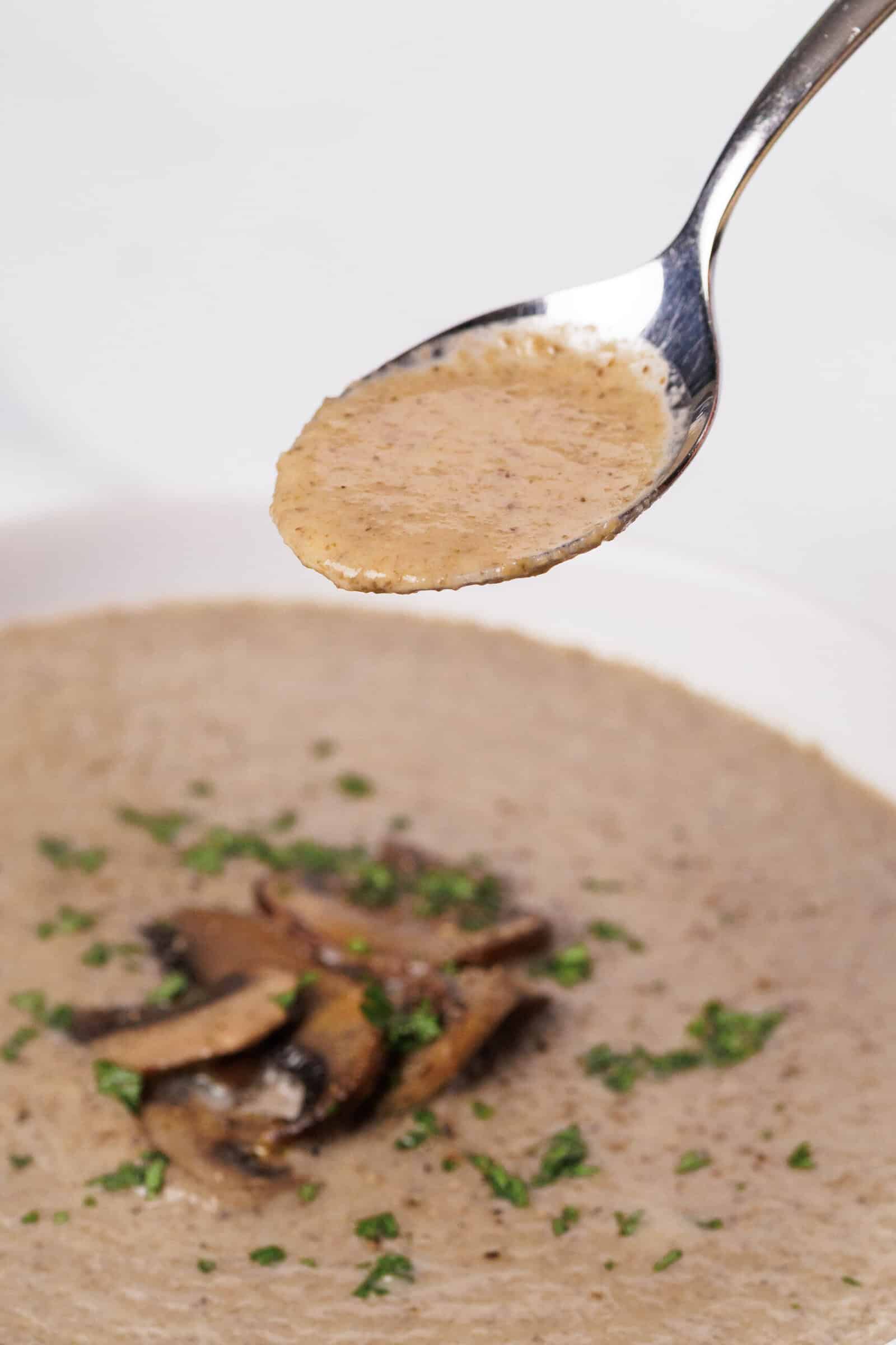 A spoonful of creamy mushroom soup is held above a bowl, which is garnished with sautéed mushroom slices and chopped fresh herbs.