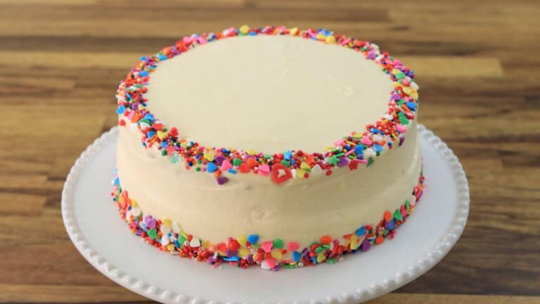 A round birthday cake with smooth white frosting is decorated with colorful sprinkles around the top and bottom edges, sitting on a white cake stand on a wooden surface.