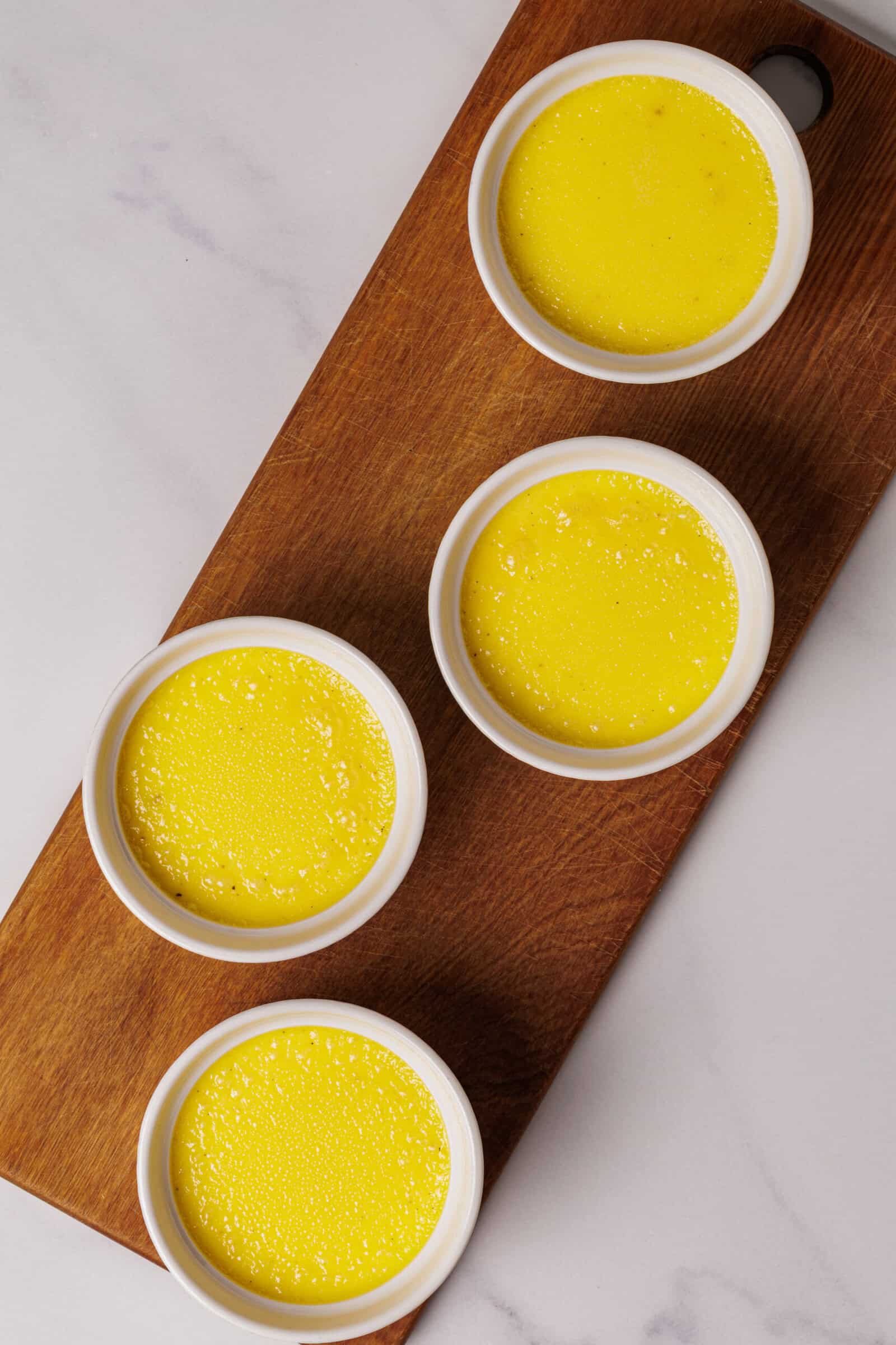 Four white ramekins filled with yellow custard are arranged on a wooden board, set against a light marble surface.
