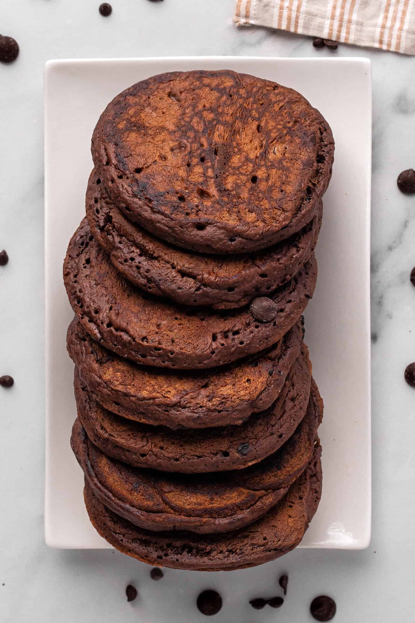 A stack of thick, fluffy chocolate pancakes sits on a rectangular white plate, with a few chocolate chips scattered around on a marble surface. A striped cloth is partially visible in the corner.