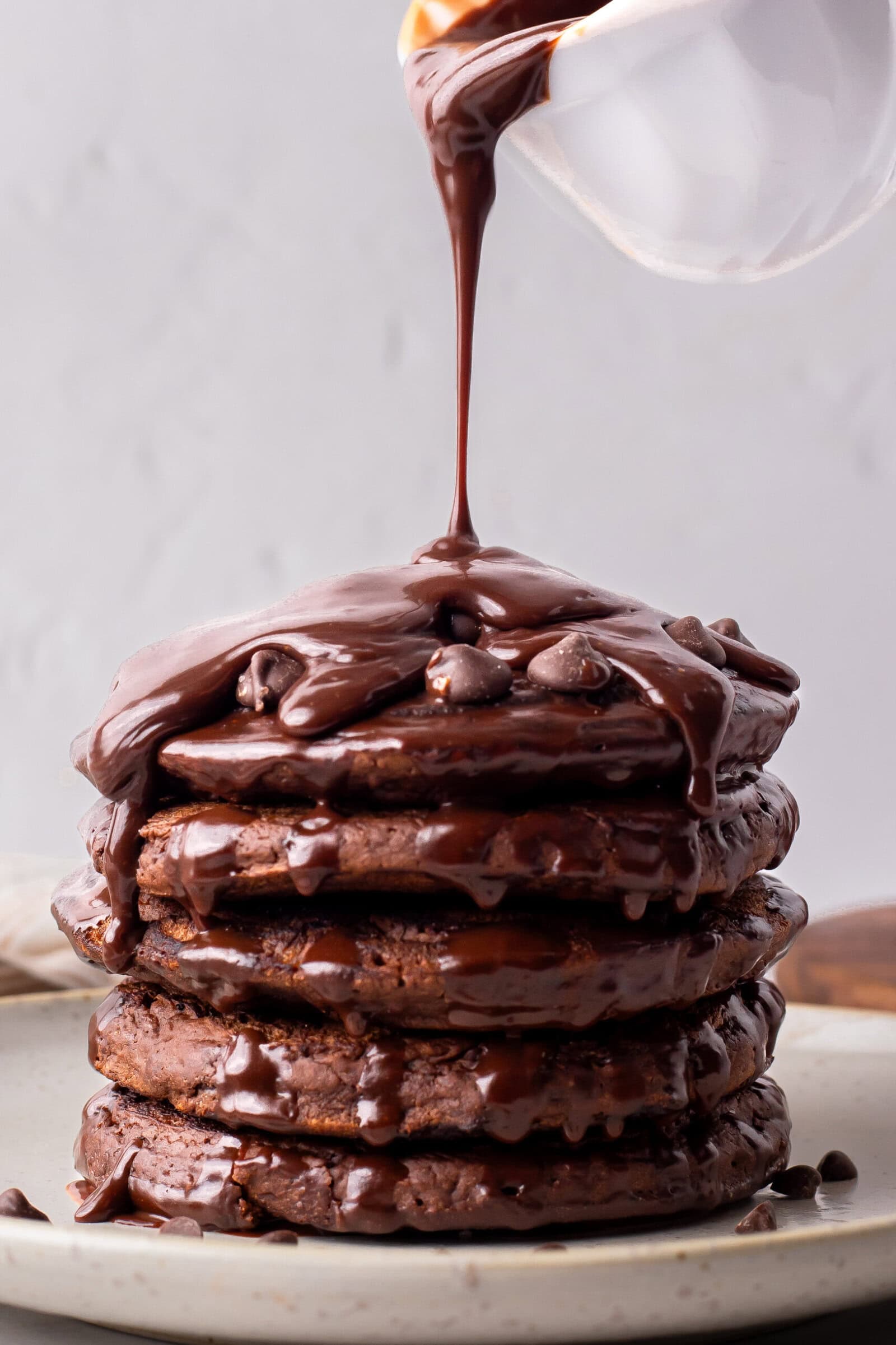 A stack of chocolate pancakes on a plate is being drizzled with rich chocolate sauce from a small white pitcher above.