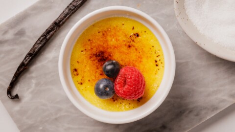Easy Crème Brûlée Recipe (4 Ingredients!) A small white ramekin of crème brûlée topped with two blueberries and a raspberry, sitting on a marble surface near a vanilla bean and a bowl of sugar.