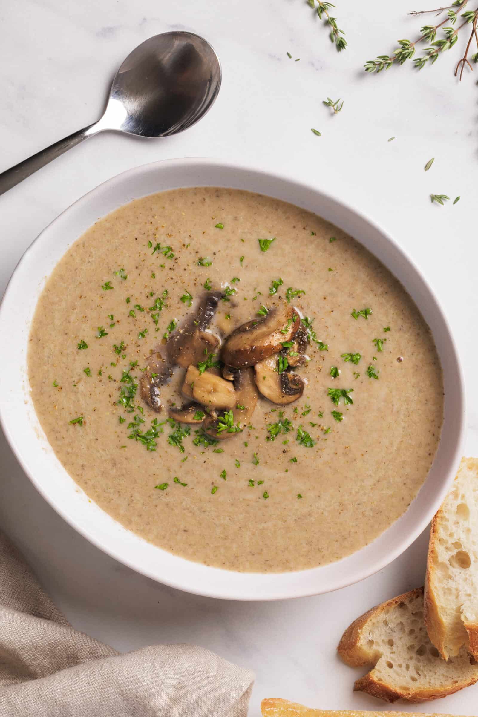 A bowl of creamy mushroom soup garnished with sautéed mushrooms and chopped herbs, served with slices of bread on the side. A spoon and sprigs of thyme rest nearby on a white marble surface.