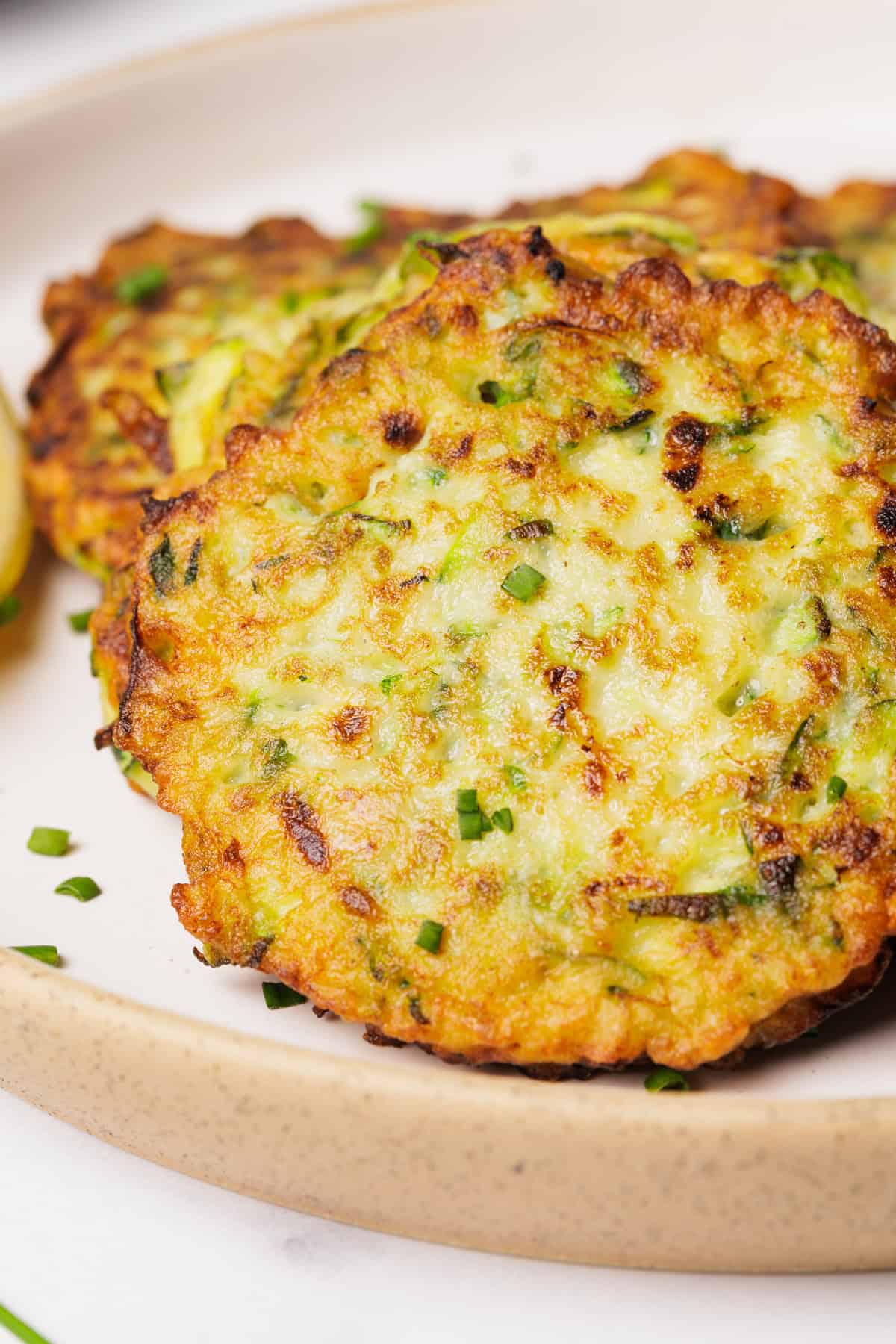 Close-up of golden-brown zucchini fritters garnished with chopped chives, served on a beige ceramic plate. The fritters have a crispy texture with visible green zucchini and herbs.