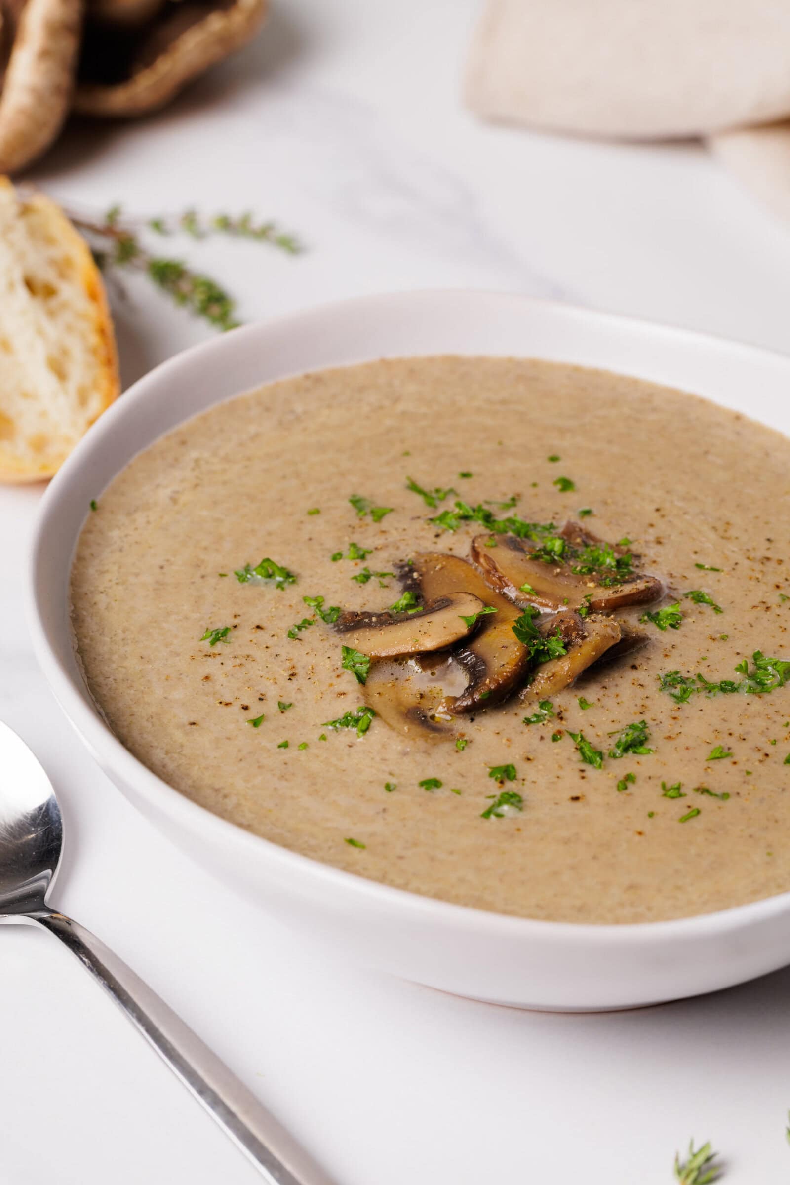 A bowl of creamy mushroom soup garnished with sautéed mushroom slices and chopped herbs, placed beside a slice of bread and a spoon on a white surface.