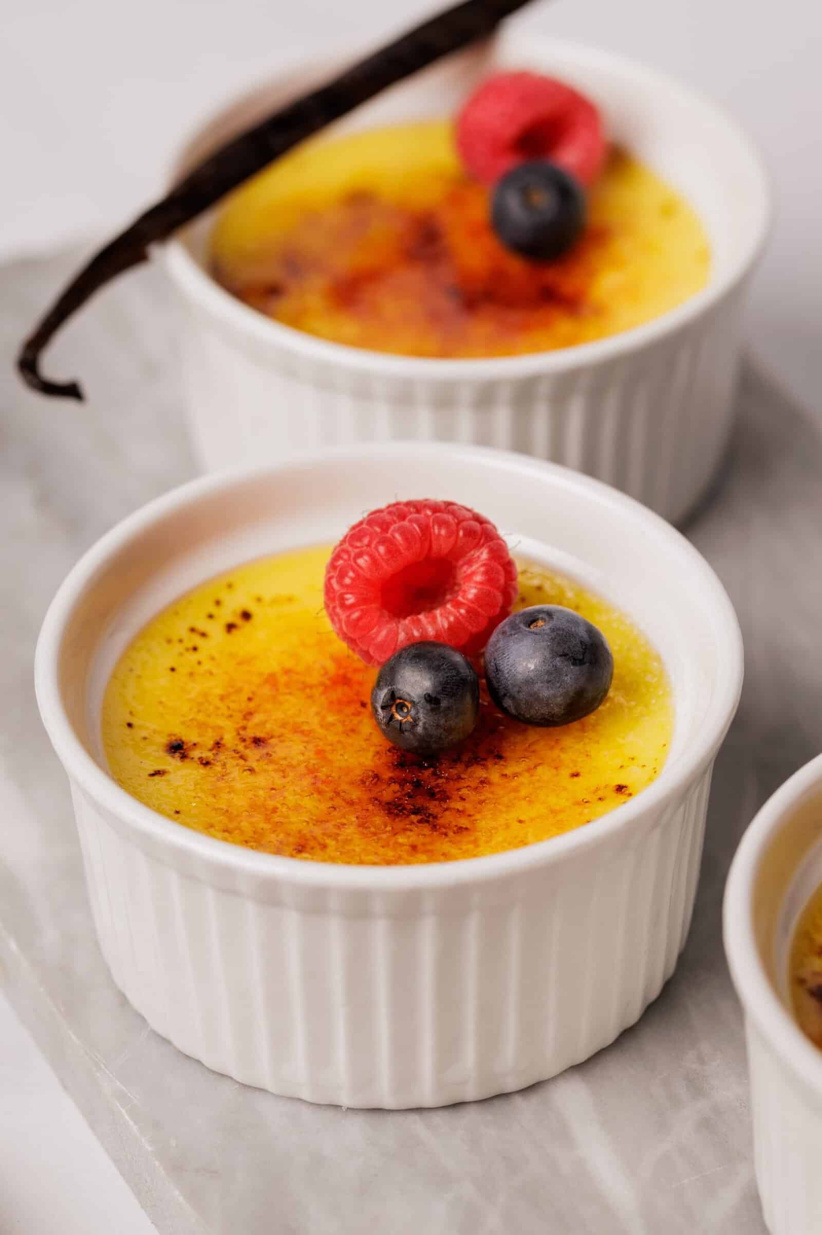 Close-up of a ramekin filled with crème brûlée, topped with a caramelized sugar crust, a raspberry, two blueberries, and a vanilla bean, with another similar dessert in the background.