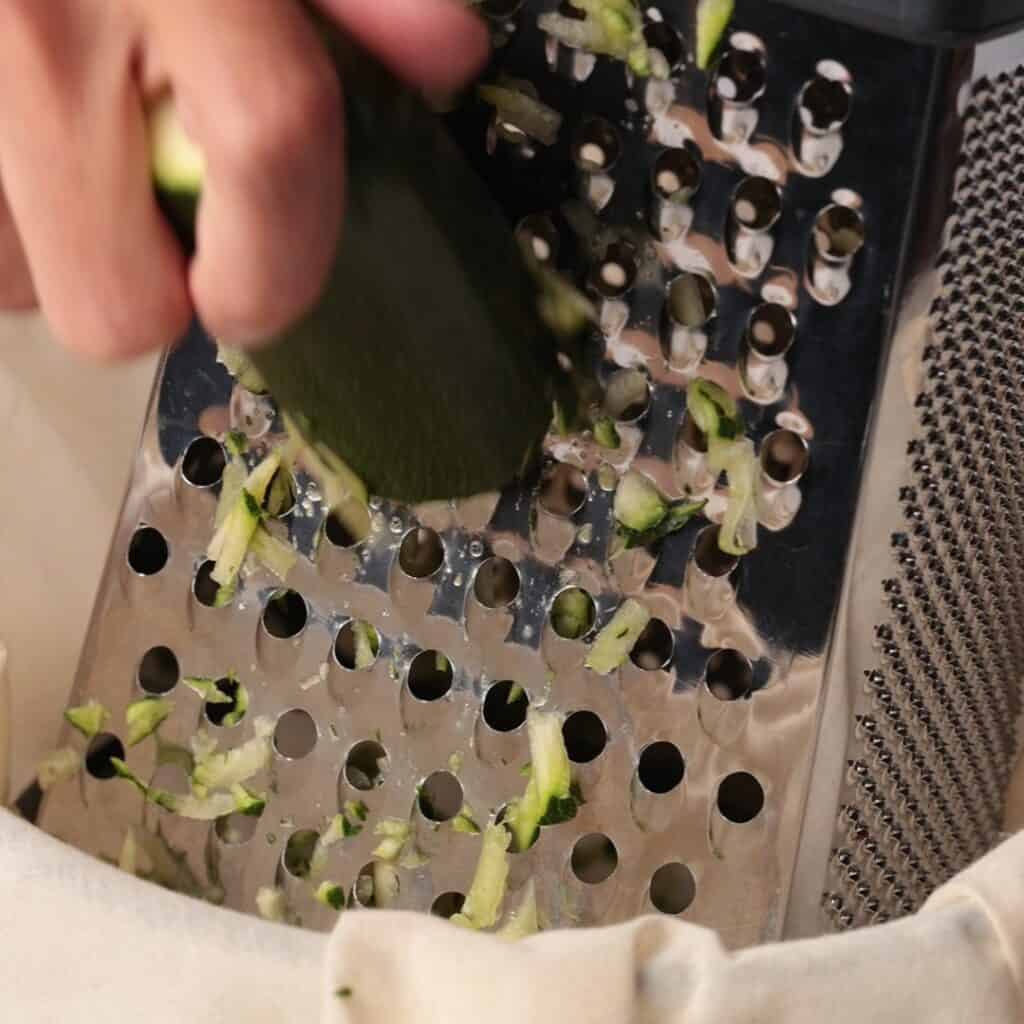 A close-up of a hand grating a zucchini using the large holes of a box grater, with zucchini shreds collecting below.