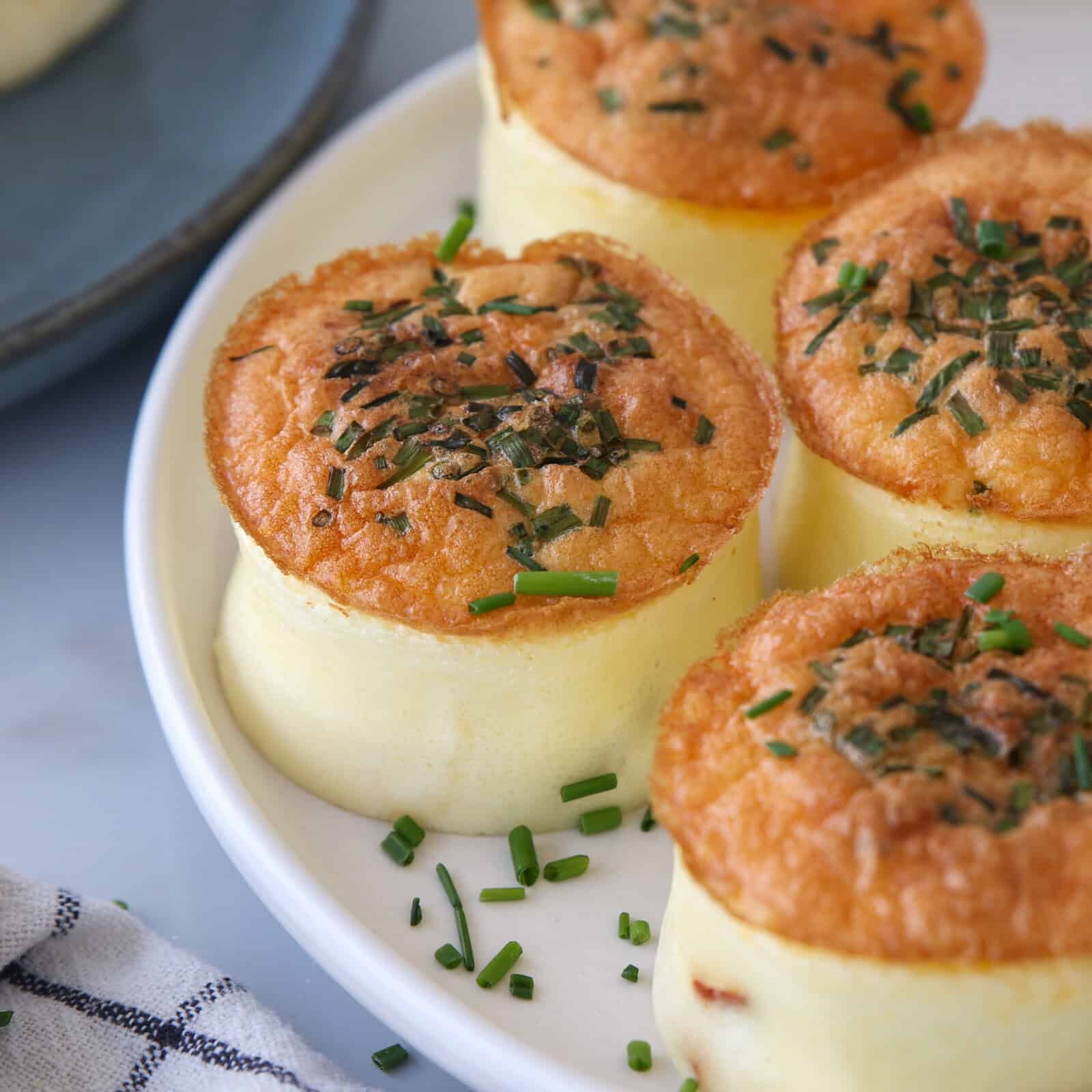 Four fluffy, golden-brown egg soufflés topped with chopped chives are arranged on a white plate. A gray plate and a checkered napkin are partially visible in the background.