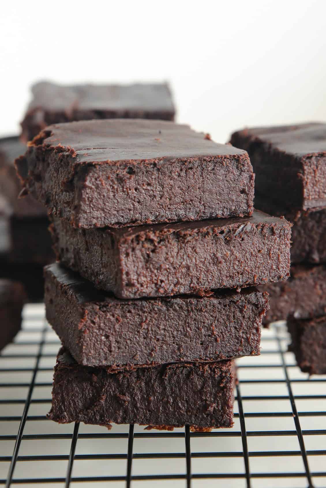 A close-up of a stack of thick, fudgy 3-ingredient chocolate brownies on a black cooling rack, with more brownies blurred in the background.