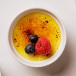 A small white ramekin filled with crème brûlée, topped with caramelized sugar, a raspberry, and two blueberries, viewed from above on a white surface.