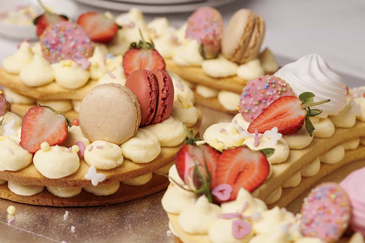 Close-up of decorative number cake topped with piped cream, sliced strawberries, macarons, meringues, and colorful sprinkles. The desserts are arranged closely together on a silver surface.