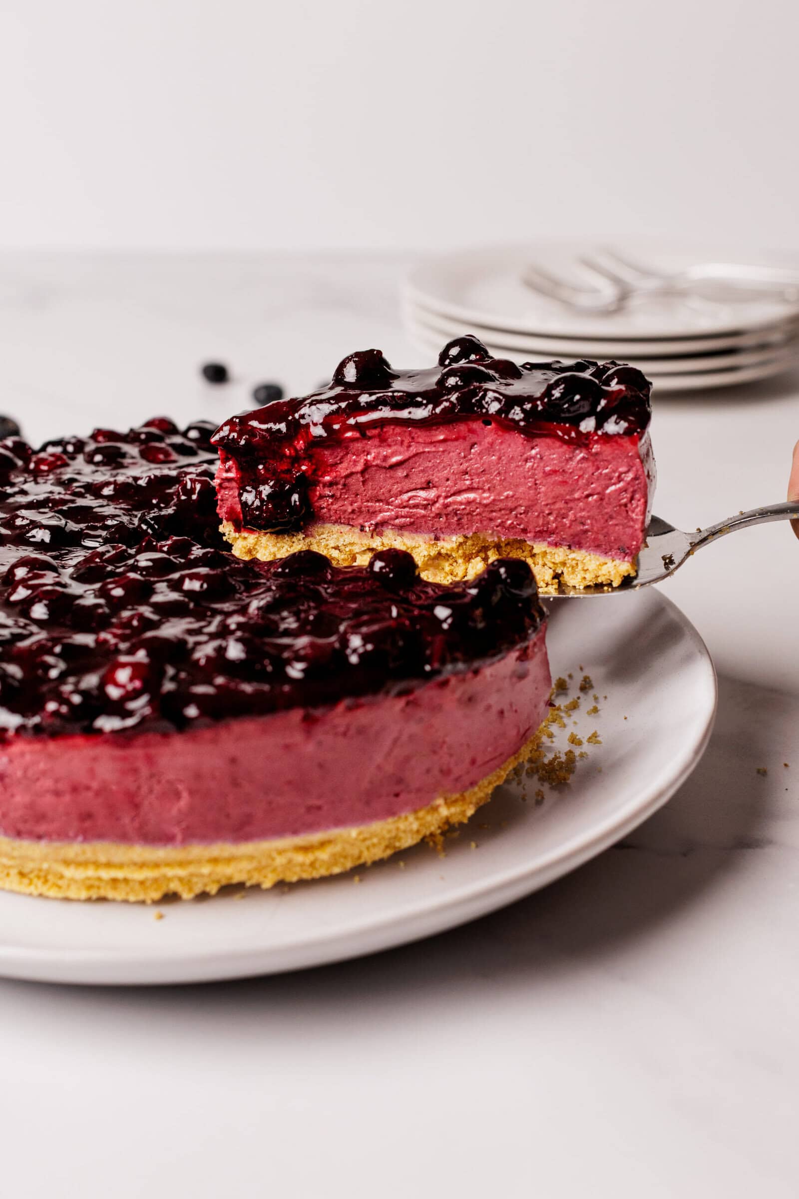 A hand lifts a slice of berry-topped cheesecake from a whole cake on a white plate, showing a crumbly base, pink filling, and glossy berry topping. Stacked plates and forks are blurred in the background.
