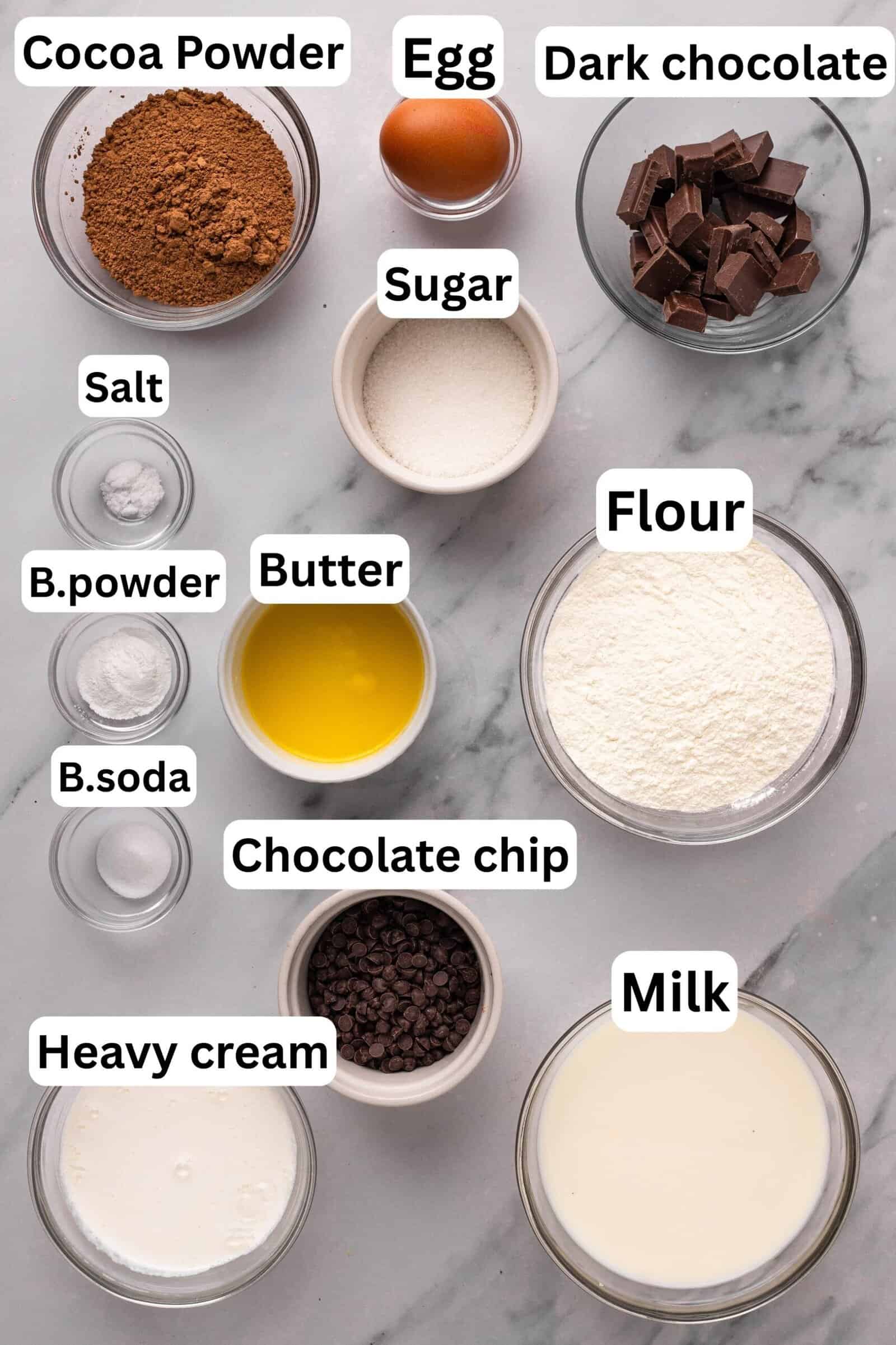A marble countertop displays labeled bowls with ingredients: cocoa powder, egg, dark chocolate, sugar, salt, baking powder, butter, flour, chocolate chips, baking soda, heavy cream, and milk.
