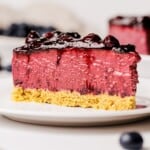 A slice of berry cheesecake with a golden crumb crust, vibrant purple filling, and glossy berry topping sits on a white plate, with blueberries blurred in the background.