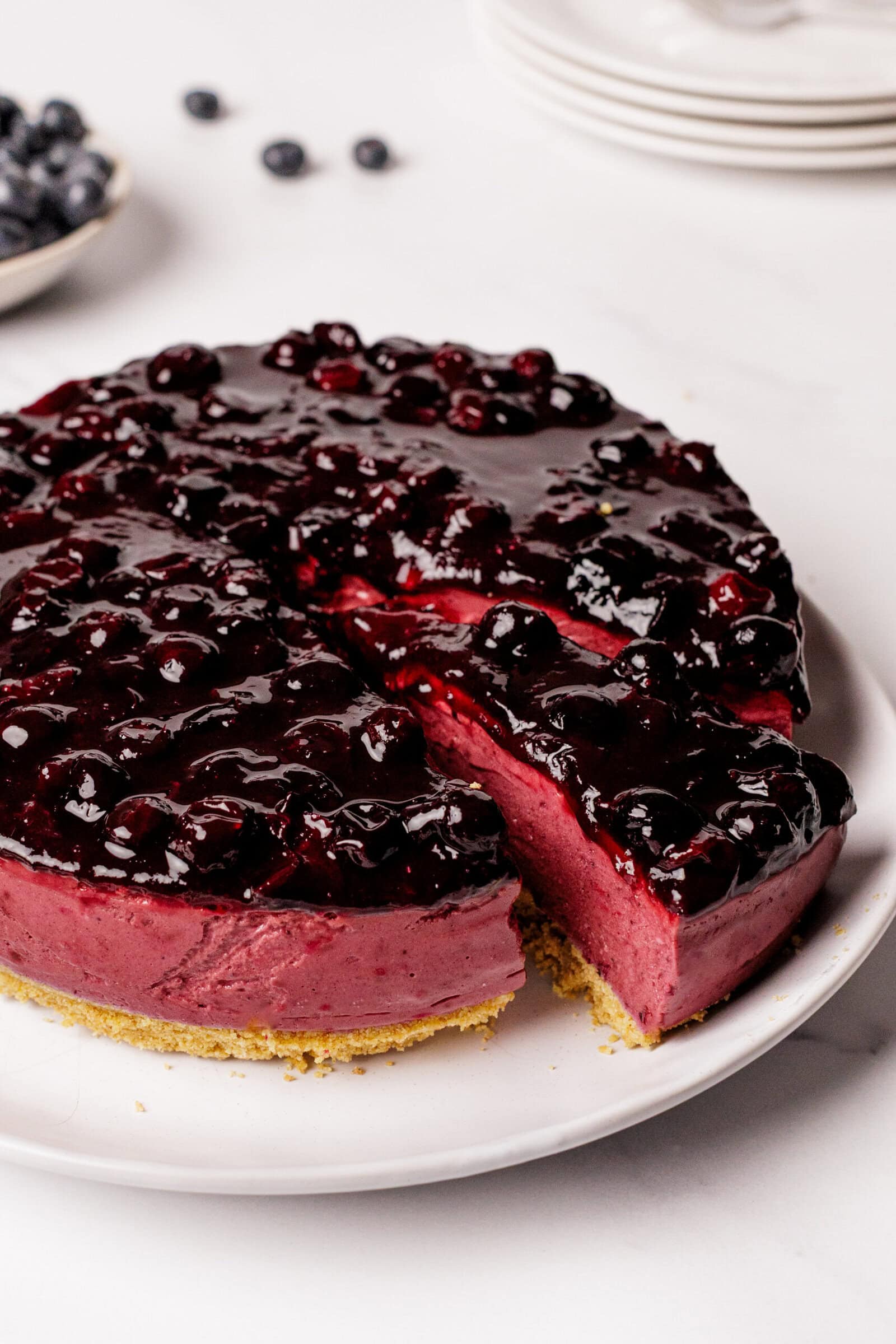A round blueberry cheesecake with a golden crust and glossy blueberry topping sits on a white plate. One slice has been cut and slightly pulled out, revealing the creamy purple filling. Plates and blueberries are visible in the background.