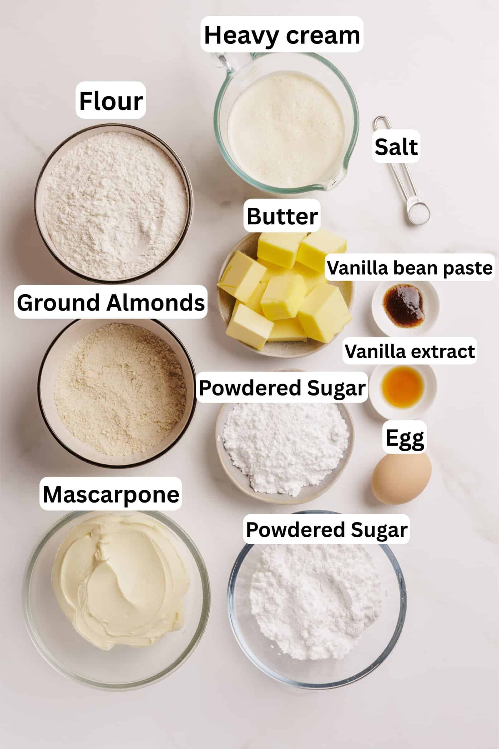 Top-down view of baking ingredients in bowls: flour, ground almonds, mascarpone, powdered sugar (in two bowls), heavy cream, cubed butter, an egg, salt, vanilla bean paste, and vanilla extract, all labeled.