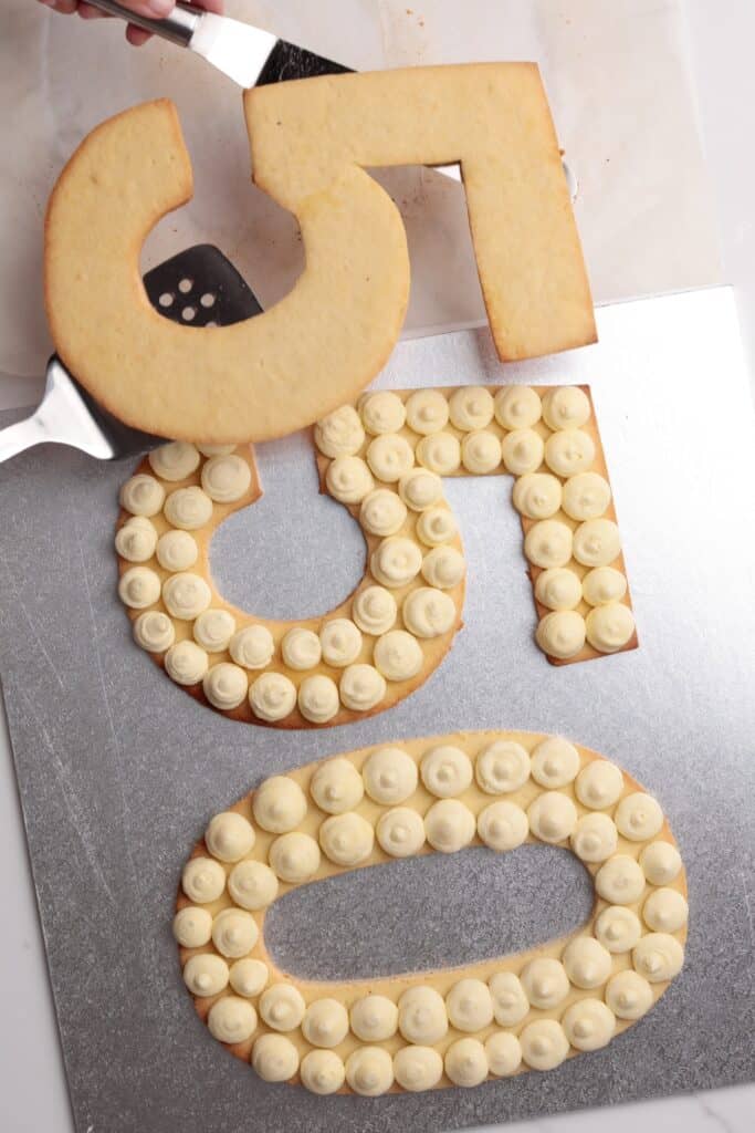 Three large cookie numbers—5, 5, and 0—are laid on a silver surface. The 5 and 0 are topped with evenly spaced dollops of white frosting. A spatula is lifting the undecorated 5.