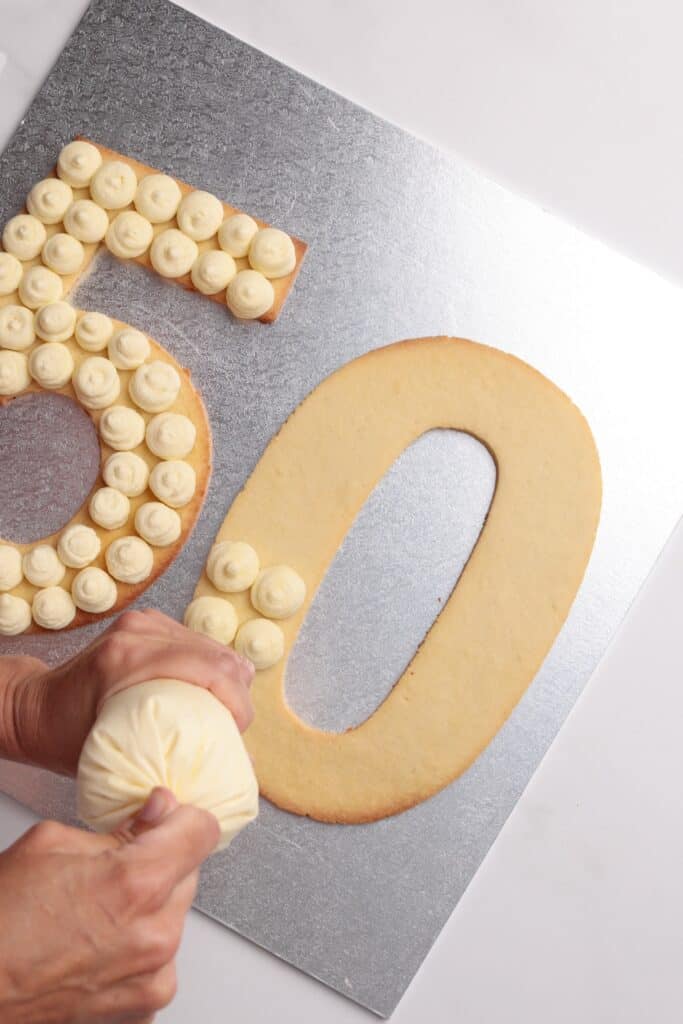 A person pipes cream onto a large, cookie-shaped number 5 on a silver board next to an undecorated number 0, forming the number 50.