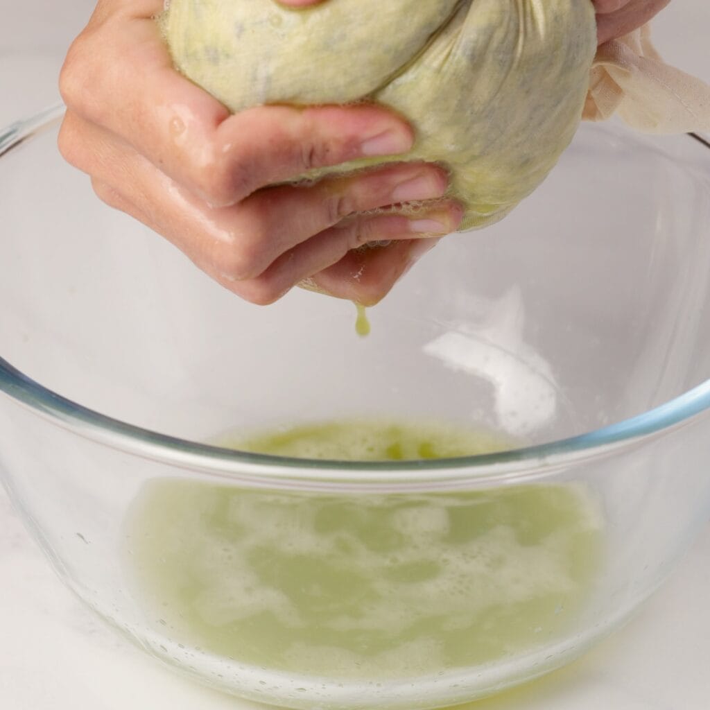 A person squeezes a cloth bundle over a glass bowl, extracting green liquid that drips into the bowl below.