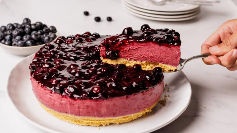 A hand lifts a slice of blueberry cheesecake with a glossy blueberry topping from a whole cheesecake on a white plate. A bowl of fresh blueberries and stacked plates are in the background.