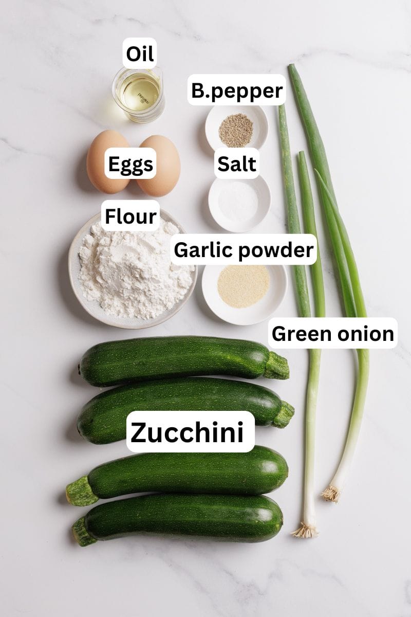 Top-down view of ingredients on a white surface: oil, eggs, flour, black pepper, salt, garlic powder, green onions, and four zucchinis. Each ingredient is labeled with text.