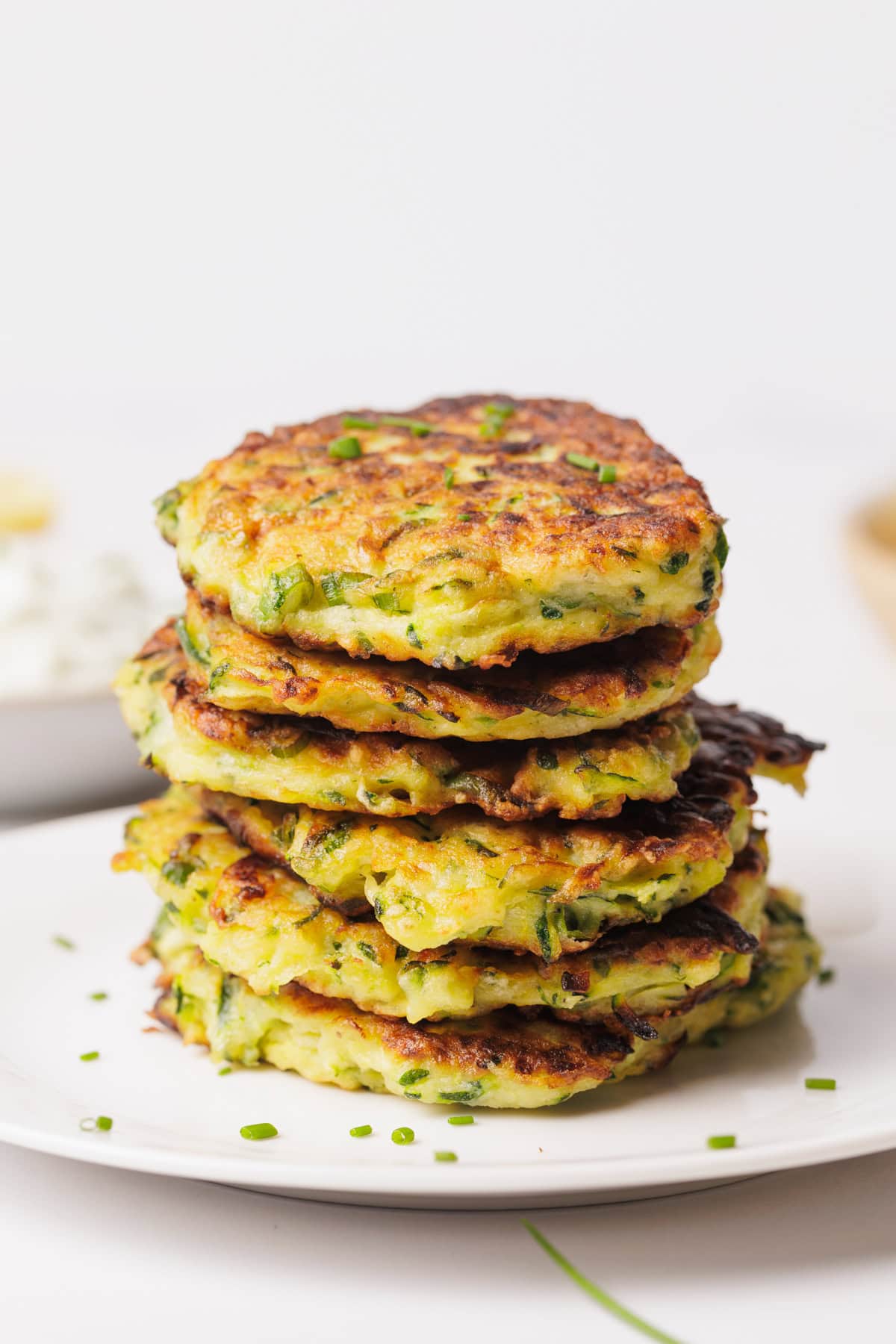 A stack of golden-brown zucchini fritters garnished with chopped herbs, arranged on a white plate against a plain white background.