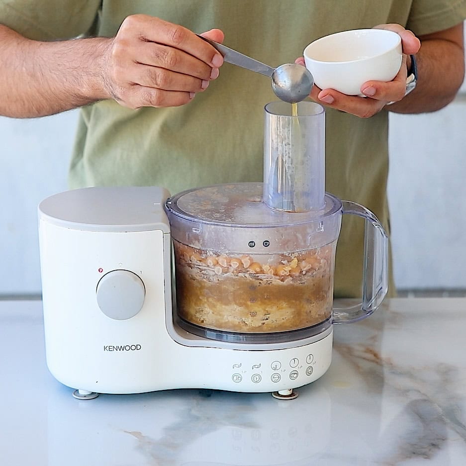 A person adds an ingredient with a spoon from a small bowl into a white Kenwood food processor containing a mixture, on a marble countertop.