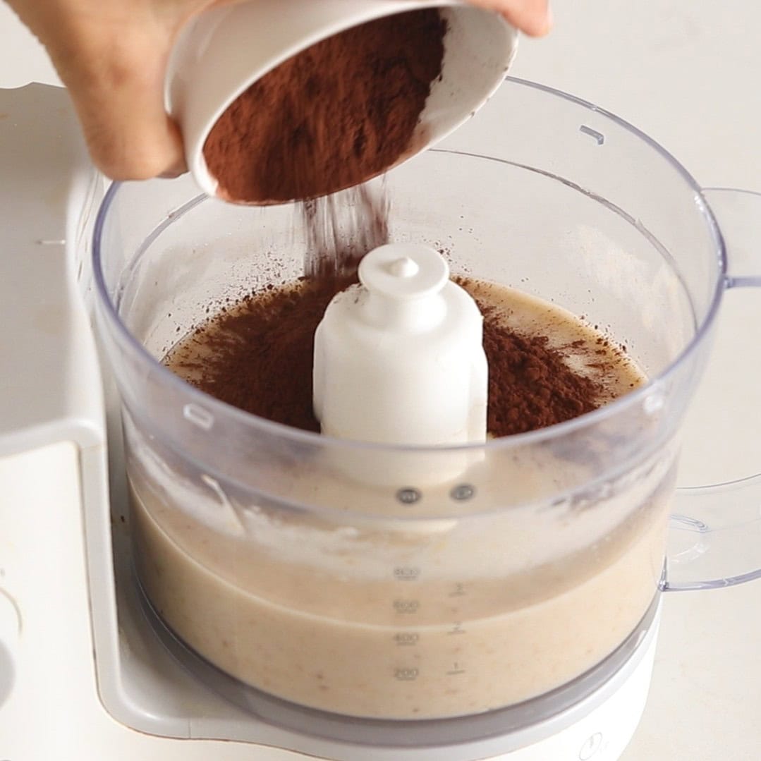 A hand pours cocoa powder from a white bowl into a food processor filled with a beige liquid mixture.