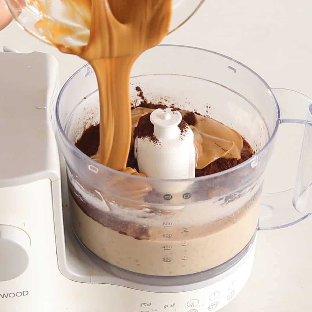 A creamy caramel-colored liquid is being poured into a food processor containing thick beige batter, cocoa powder, and peanut butter. The food processor sits on a white surface.
