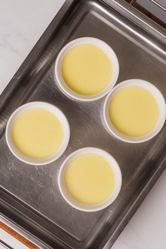 Four white ramekins filled with a pale yellow custard sit in a metal baking dish, ready to be baked in a water bath. The scene is viewed from above on a light countertop.