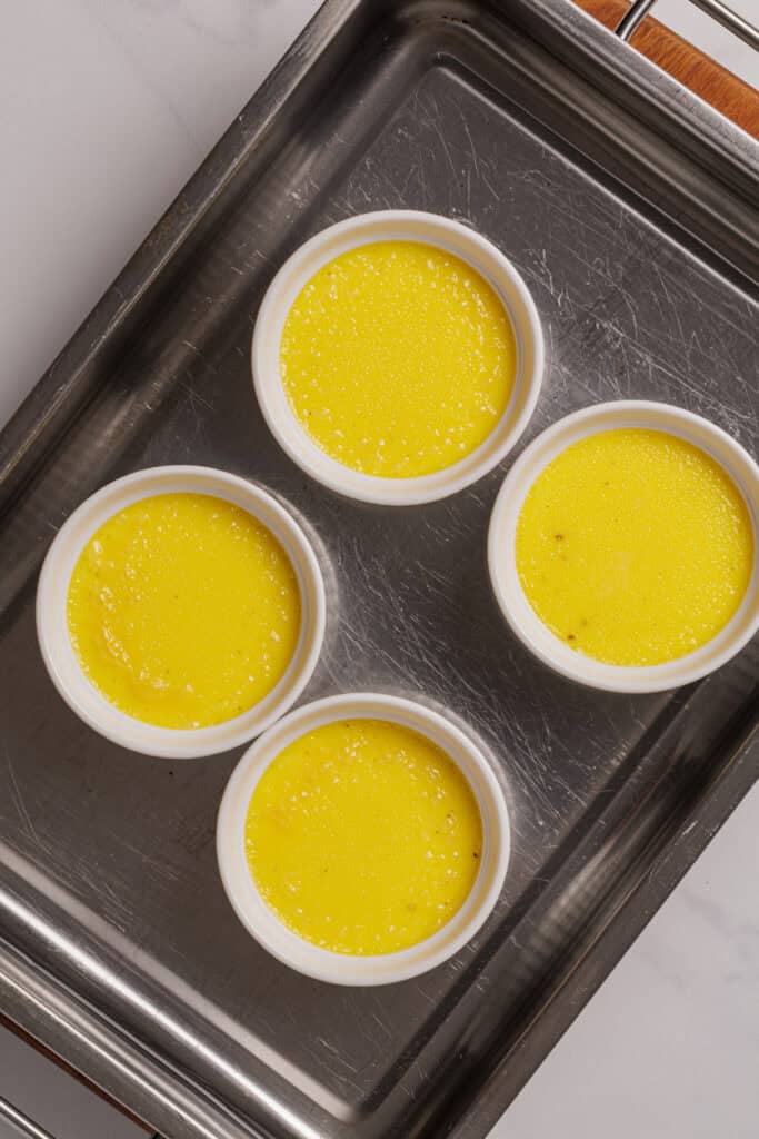 Four white ramekins filled with yellow custard are arranged in a metal baking pan, ready for baking. The surface of the custard is smooth with tiny bubbles, and the pan sits on a light-colored countertop.